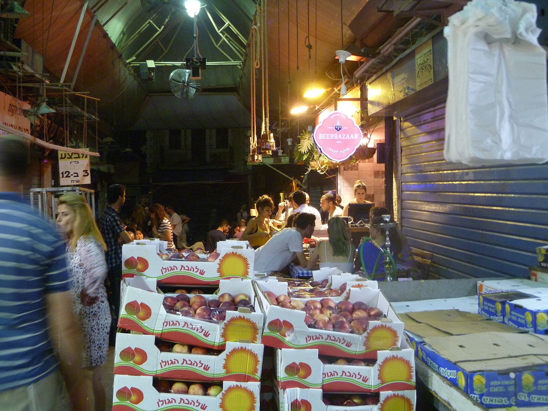 Young people drink at the Beer Bazaar bar in Mahane Yehuda Market at night, beside a closed blue shutter and stacked crates of nectarines in the foreground