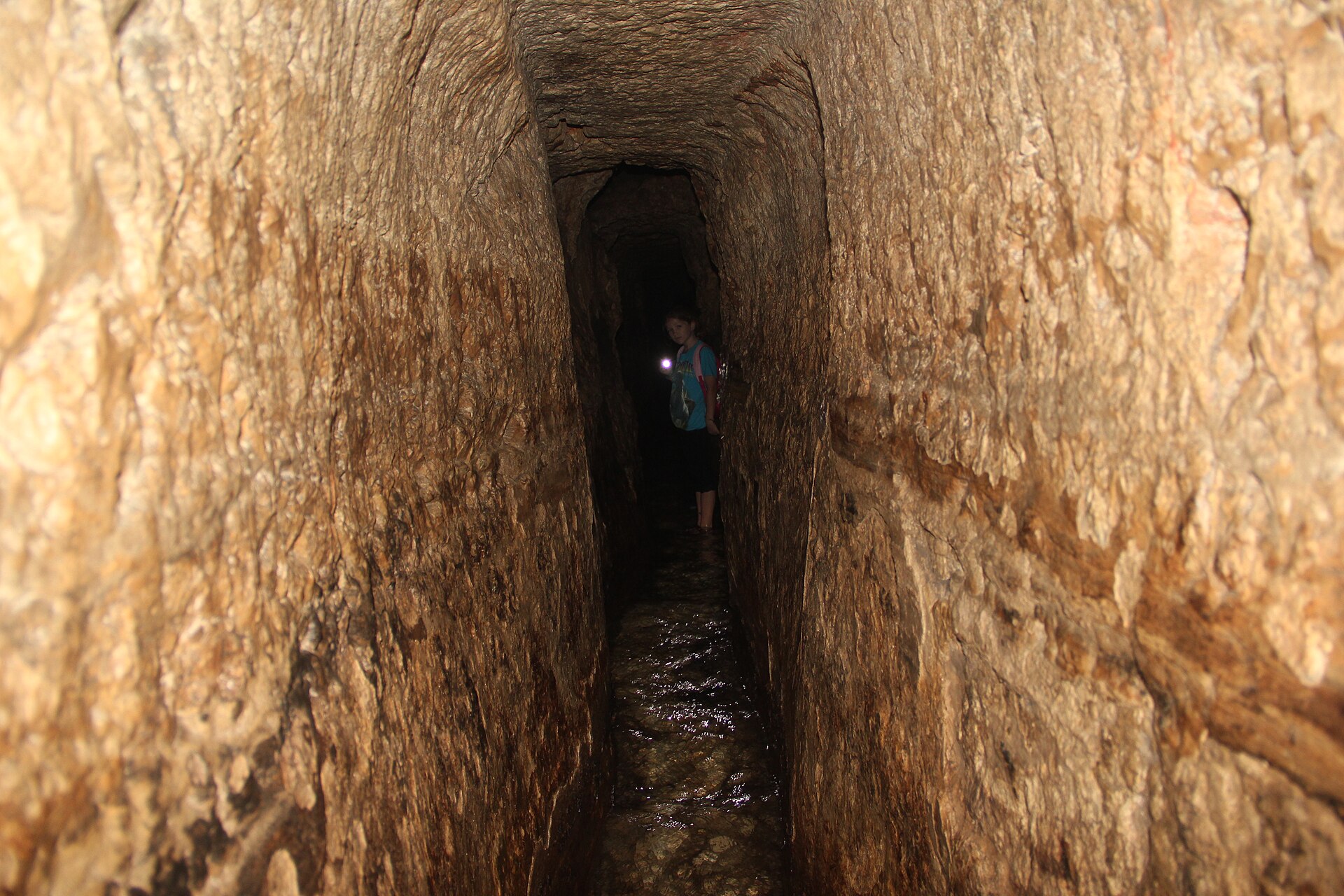 A young visitor holding a torch stands in shallow water midway along the tall, narrow rock-cut passage of Hezekiah's Tunnel