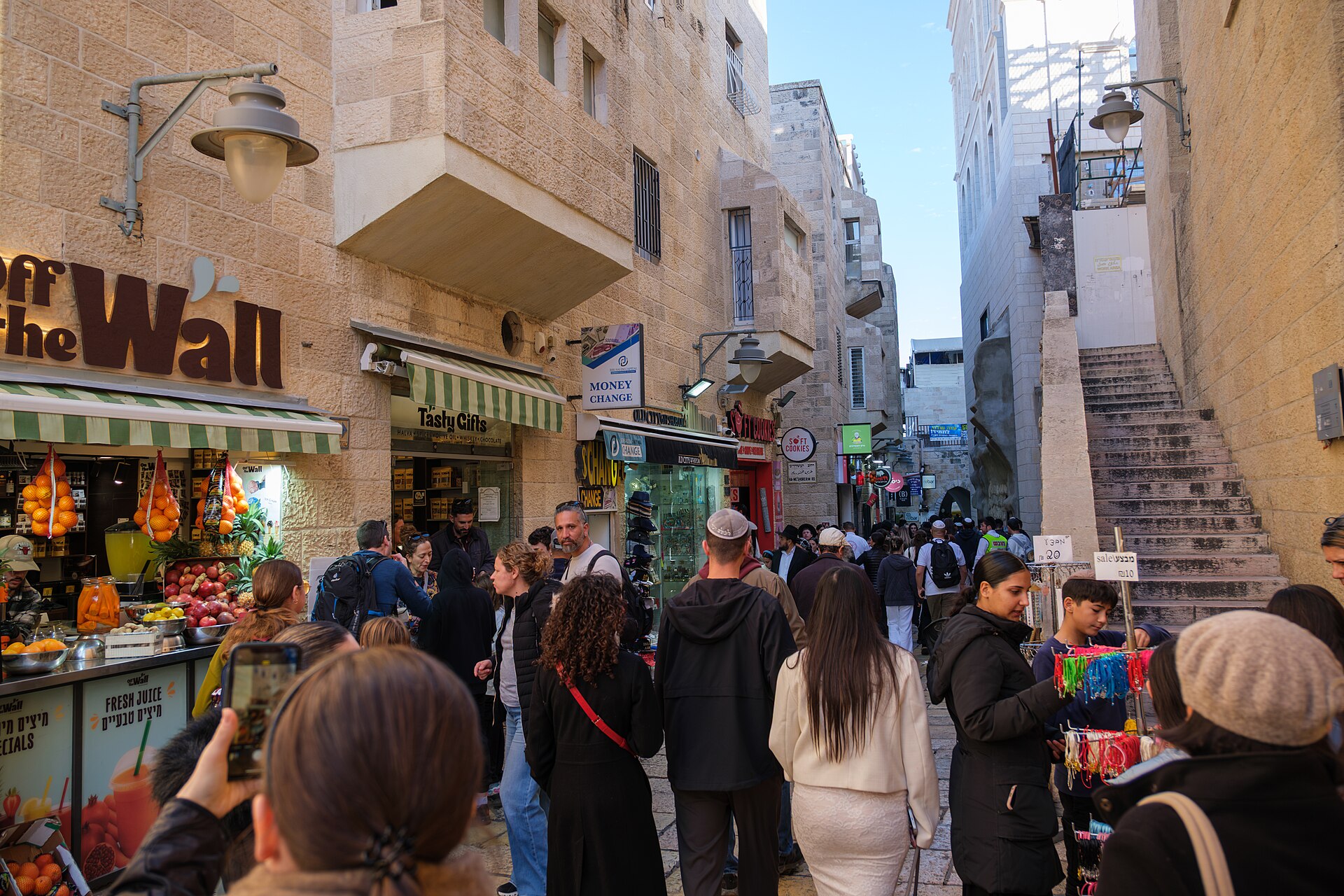 A young visitor photographs a busy Jewish Quarter lane on her phone, past a fresh juice stall, gift shops and a boy browsing a bracelet stand
