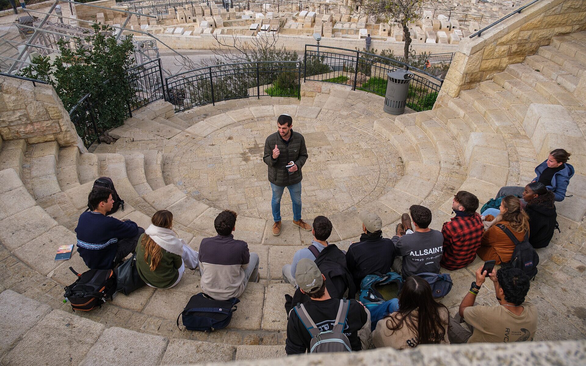 Young visitors with backpacks sit on curved stone steps listening to a guide at a Mount of Olives lookout, with the hillside Jewish cemetery below