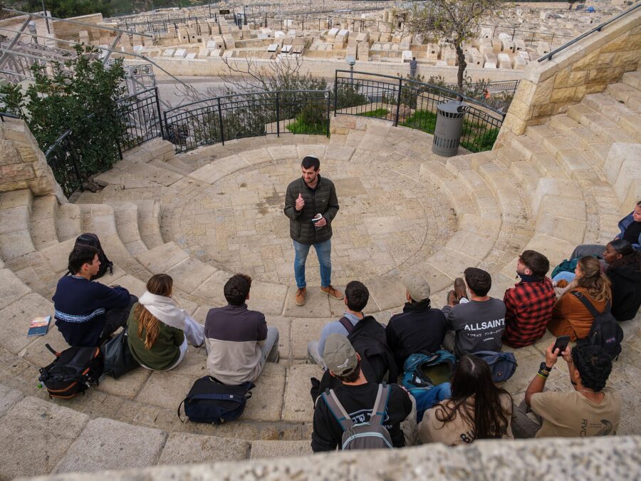 Young visitors with backpacks sit on curved stone steps listening to a guide at a Mount of Olives lookout, with the hillside Jewish cemetery below