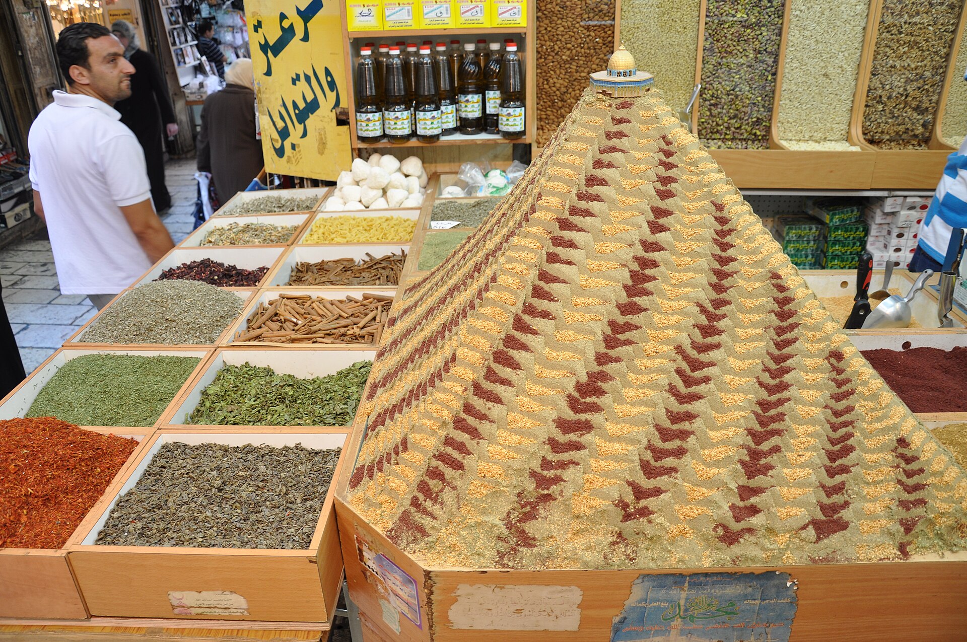 A za'atar pyramid topped by a Dome of the Rock model and trays of cinnamon, hibiscus and dried herbs at a spice shop opening onto an Old City souk lane