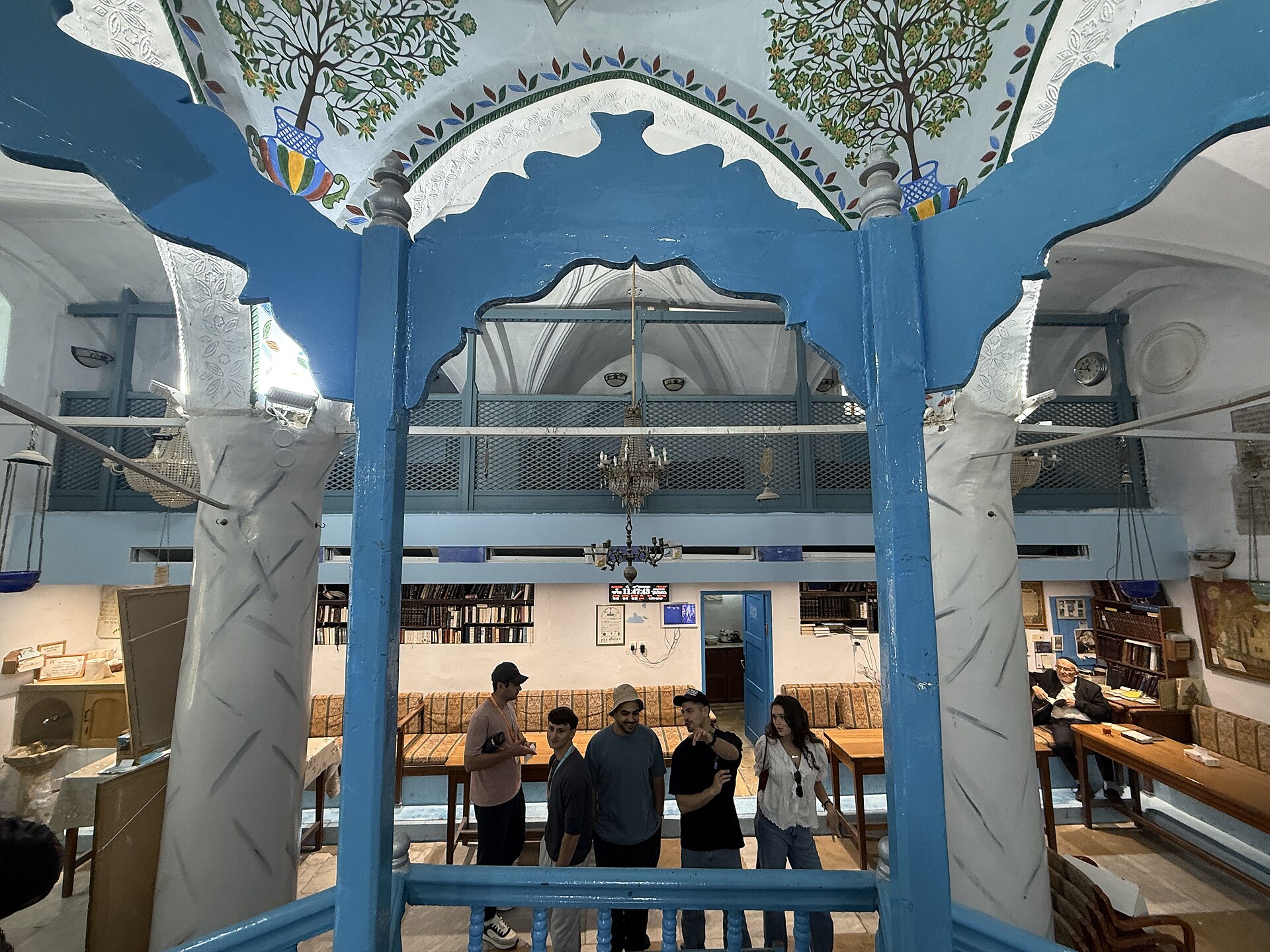 Interior of the Abuhav Synagogue in Safed, with blue-painted arches and columns, a central raised platform and visitors standing among wooden benches