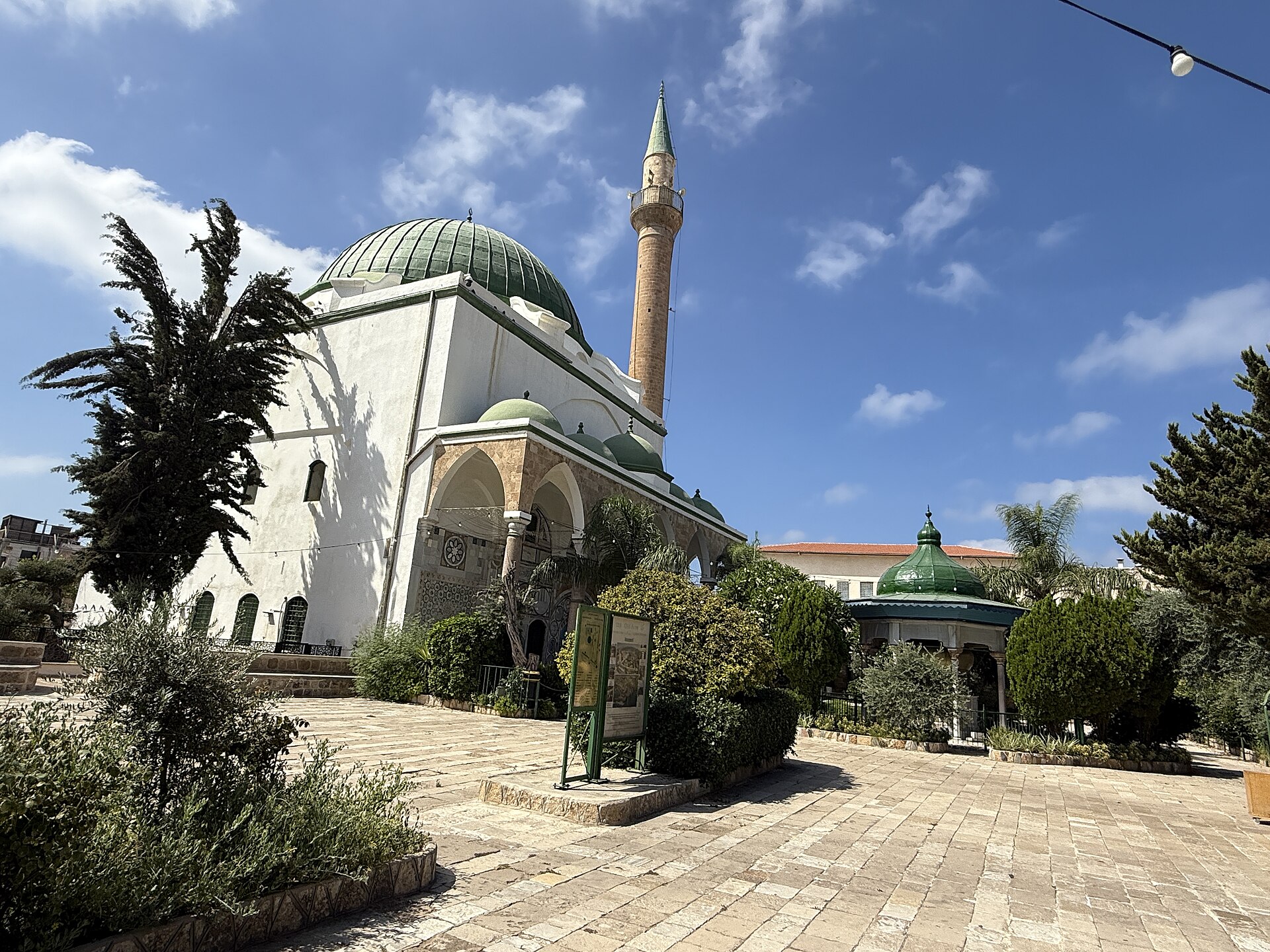 A large white mosque with a green dome and a tall slender minaret, seen across a paved courtyard with palms and trees under a blue sky
