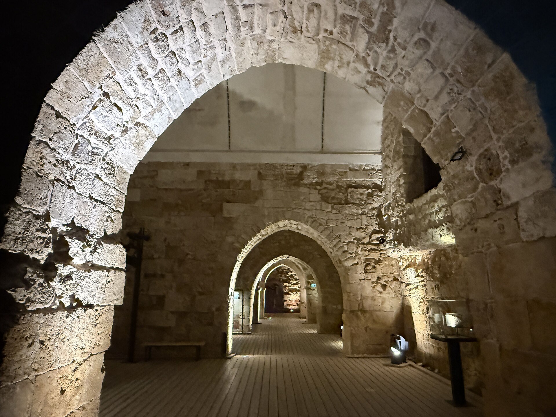A long vaulted stone hall with pointed arches receding into the distance, lit from below, with a wooden walkway along the floor