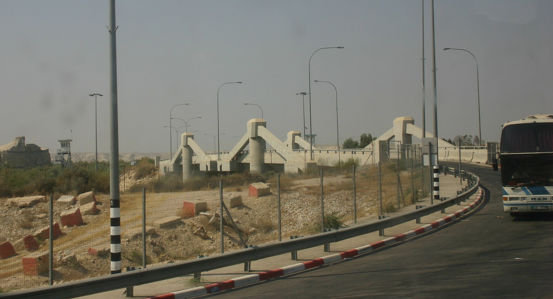 The concrete Allenby / King Hussein Bridge across the Jordan Valley seen from the curving approach road, with a watchtower, fencing and a bus