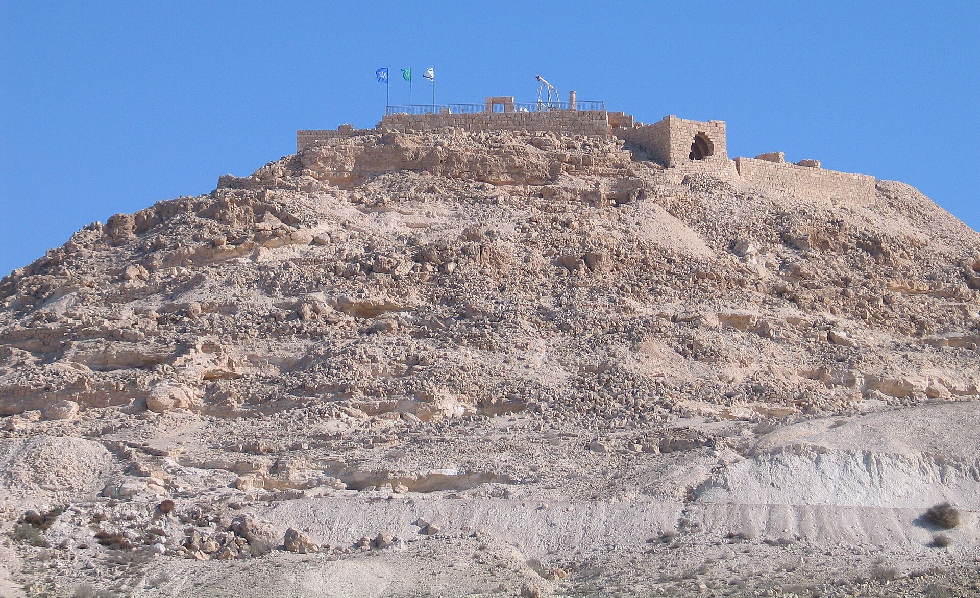 Ancient stone ruins with walls and arches crowning the top of a rocky desert hill under a clear blue sky