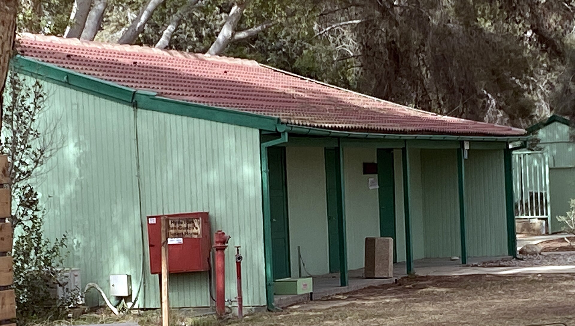 A long single-storey wooden hut painted pale green with a red tiled roof, set among trees, with a red information board in front