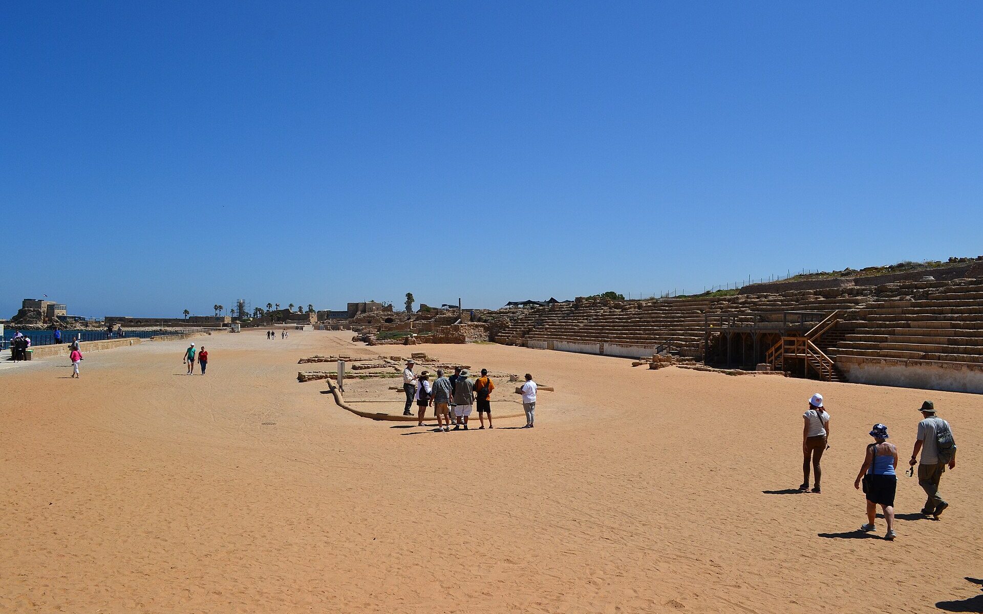 Visitors walking across the sandy track of the ancient hippodrome at Caesarea, with rows of stone seating on the right and the sea and ruins on the horizon