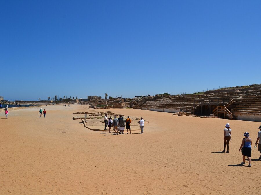 Visitors walking across the sandy track of the ancient hippodrome at Caesarea, with rows of stone seating on the right and the sea and ruins on the horizon