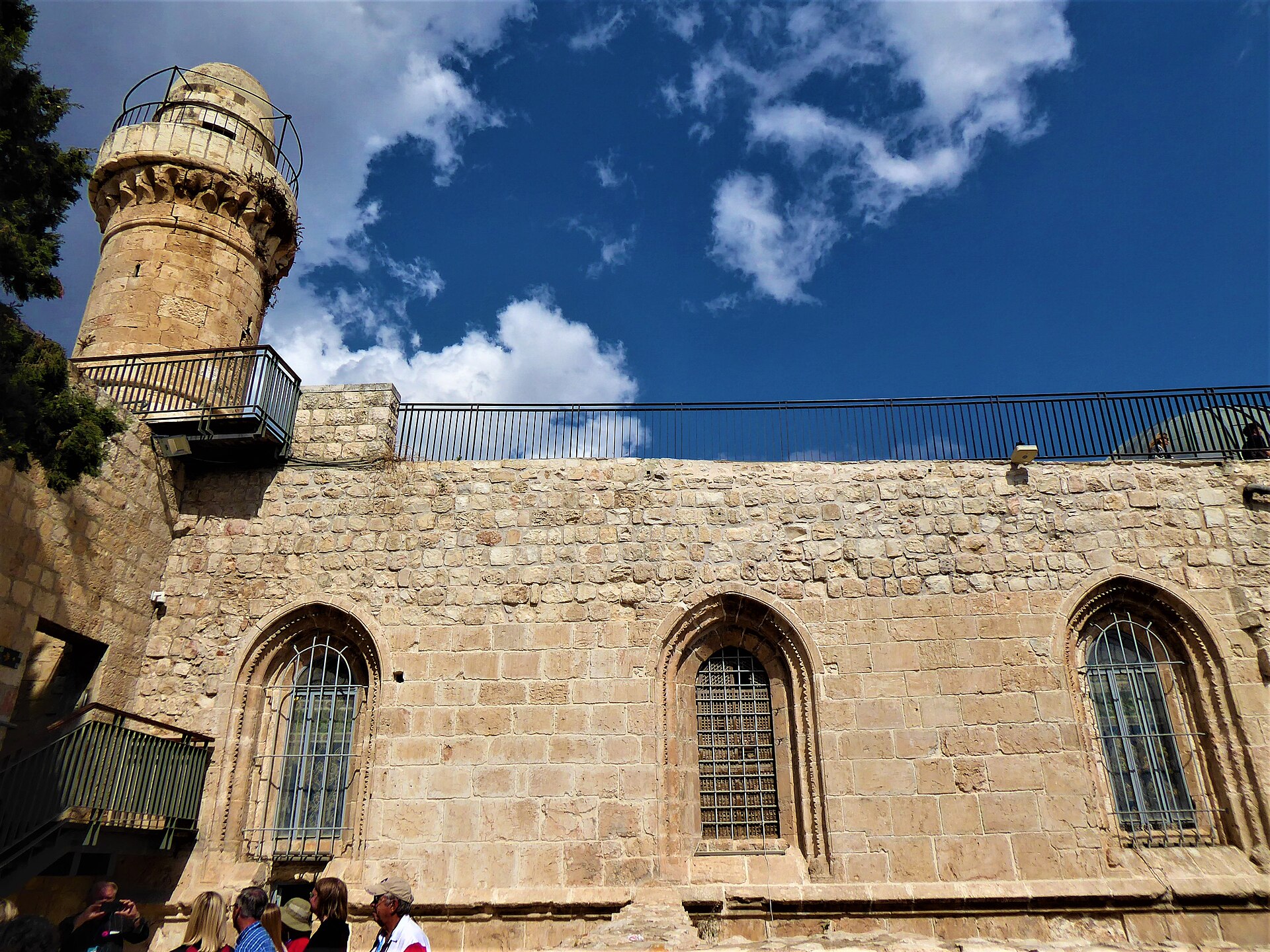 The long stone wall of a medieval building with three tall pointed windows, a railing along the roof and a stone minaret rising above it at the left under a blue sky with clouds