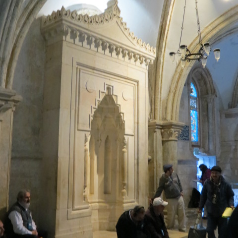A tall carved stone prayer niche set into the wall of a vaulted stone hall, with visitors sitting and standing around it and Gothic arches above