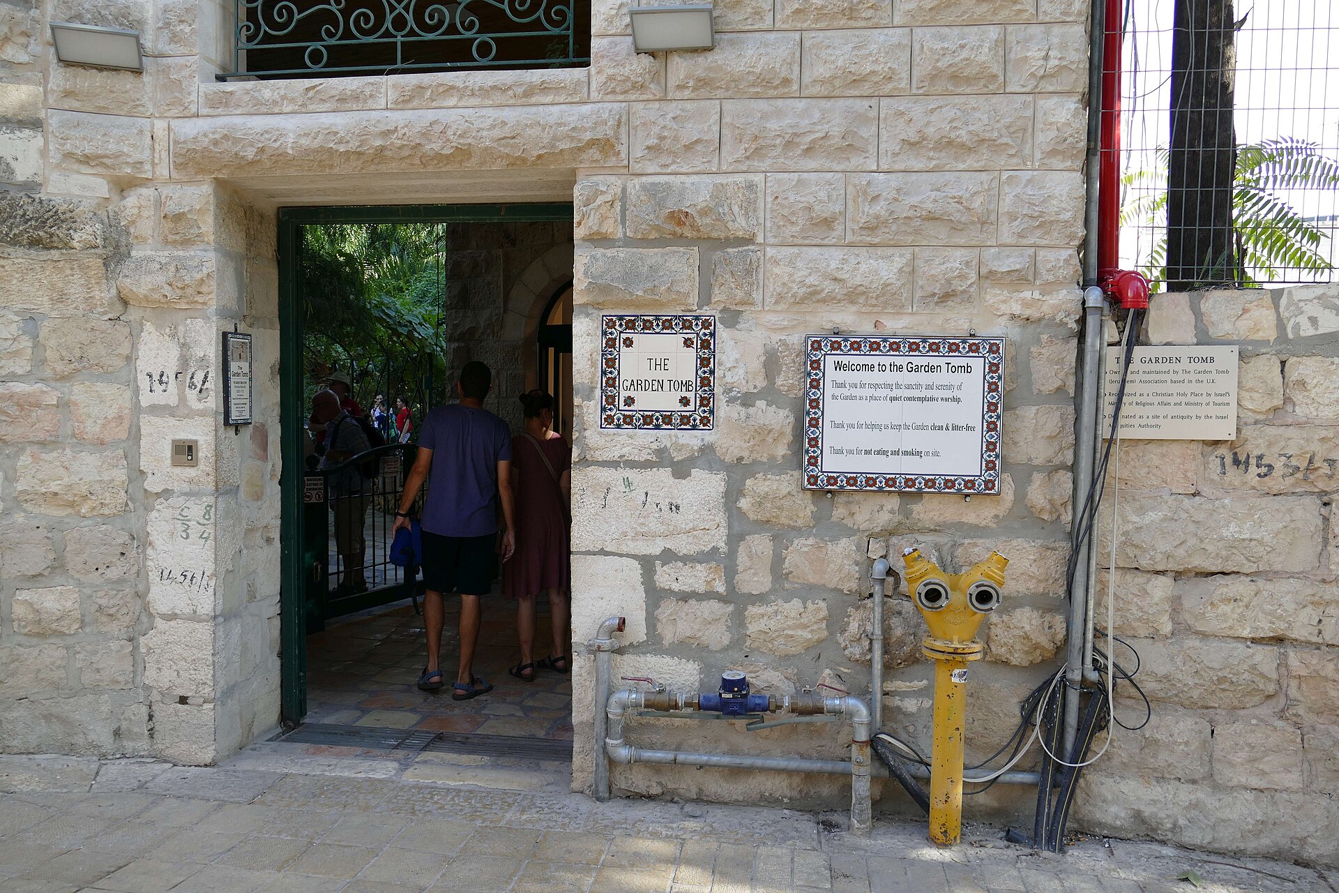 A stone gateway in a pale stone wall with a green door, a tiled sign reading The Garden Tomb, and visitors walking through into a shaded garden