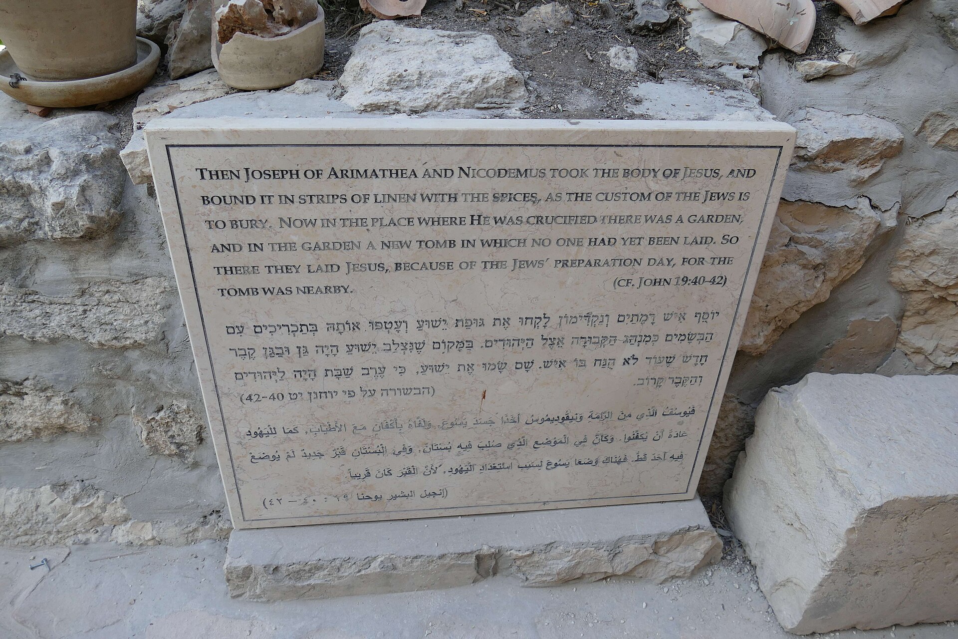 A stone plaque leaning against rough rock, engraved with a passage from the Gospel of John in English, Hebrew and Arabic, beside a pot plant