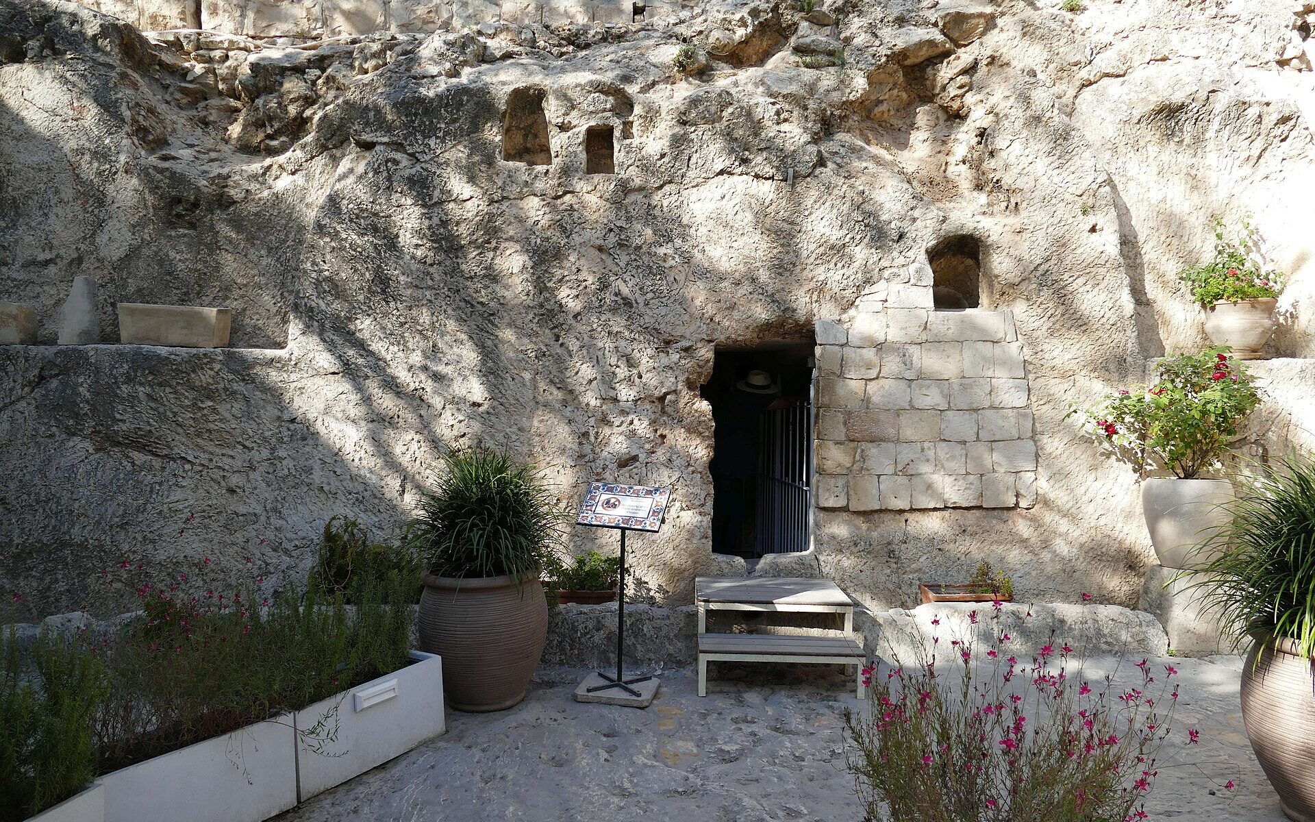 A rock face with a small dark doorway cut into it, partly walled with stone blocks, with potted plants and flowers in front