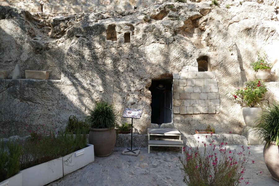 A rock face with a small dark doorway cut into it, partly walled with stone blocks, with potted plants and flowers in front