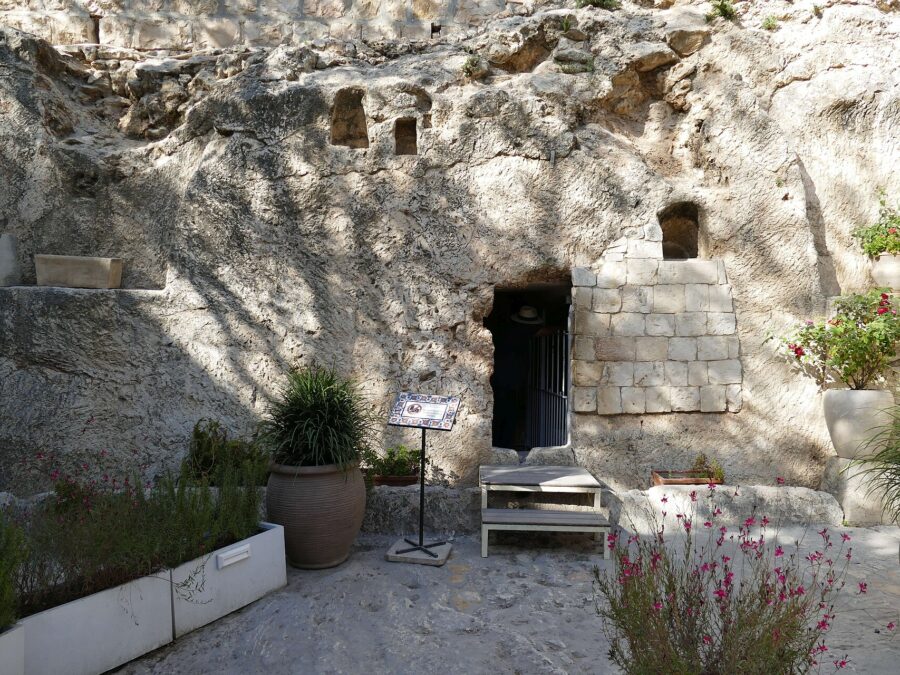 A rock face with a small dark doorway cut into it, partly walled with stone blocks, with potted plants and flowers in front