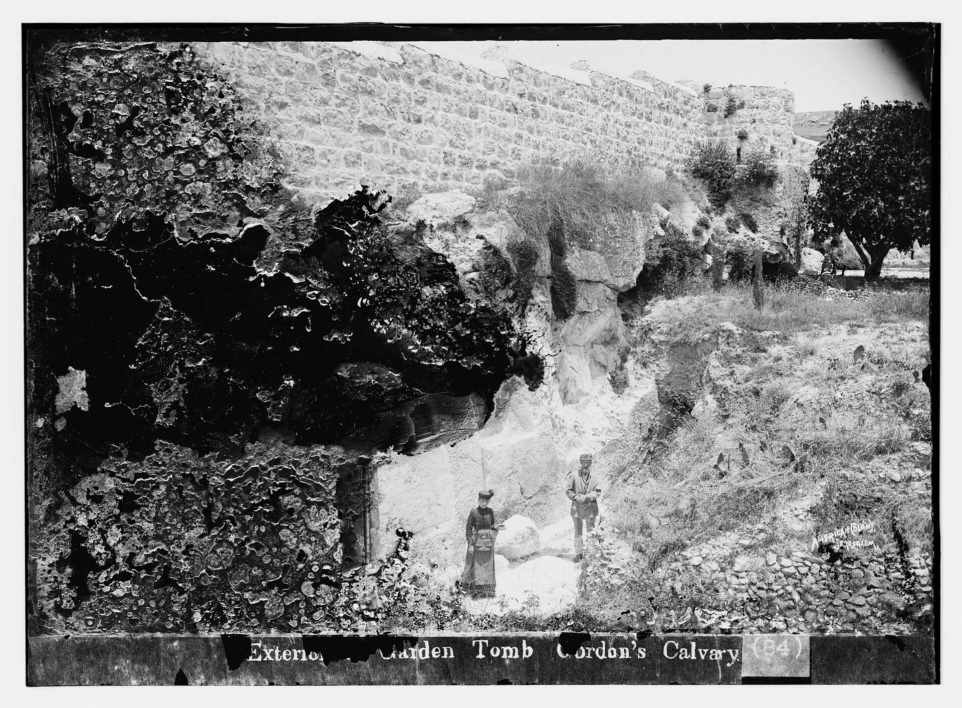 An old black-and-white photograph of a rock face with dark hollows and overgrown vegetation, with two small figures standing in front, captioned at the bottom Exterior Garden Tomb Gordon's Calvary