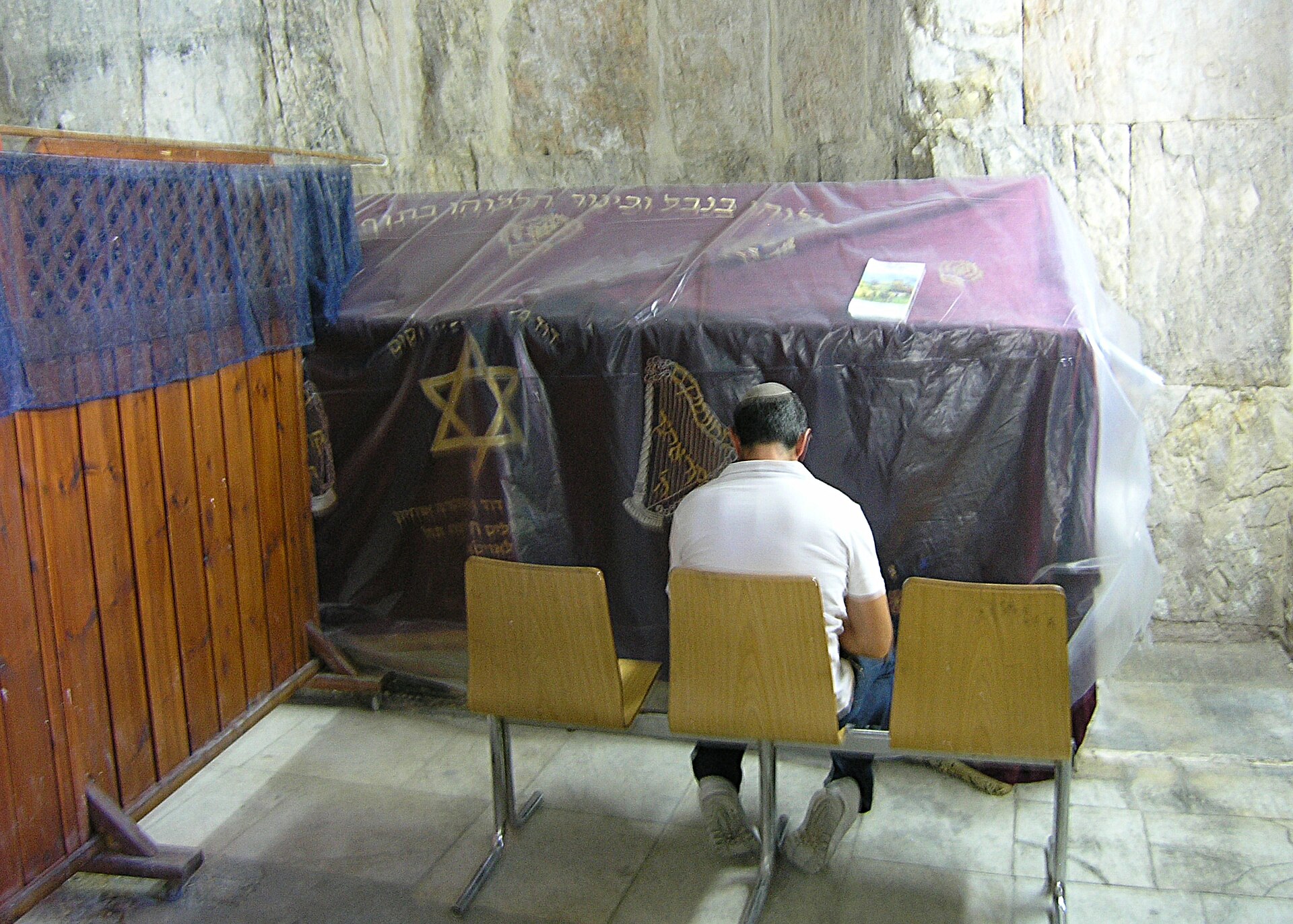 A man in a white skullcap sitting on chairs in front of a large cenotaph covered with a dark cloth embroidered with a Star of David, behind a wooden partition