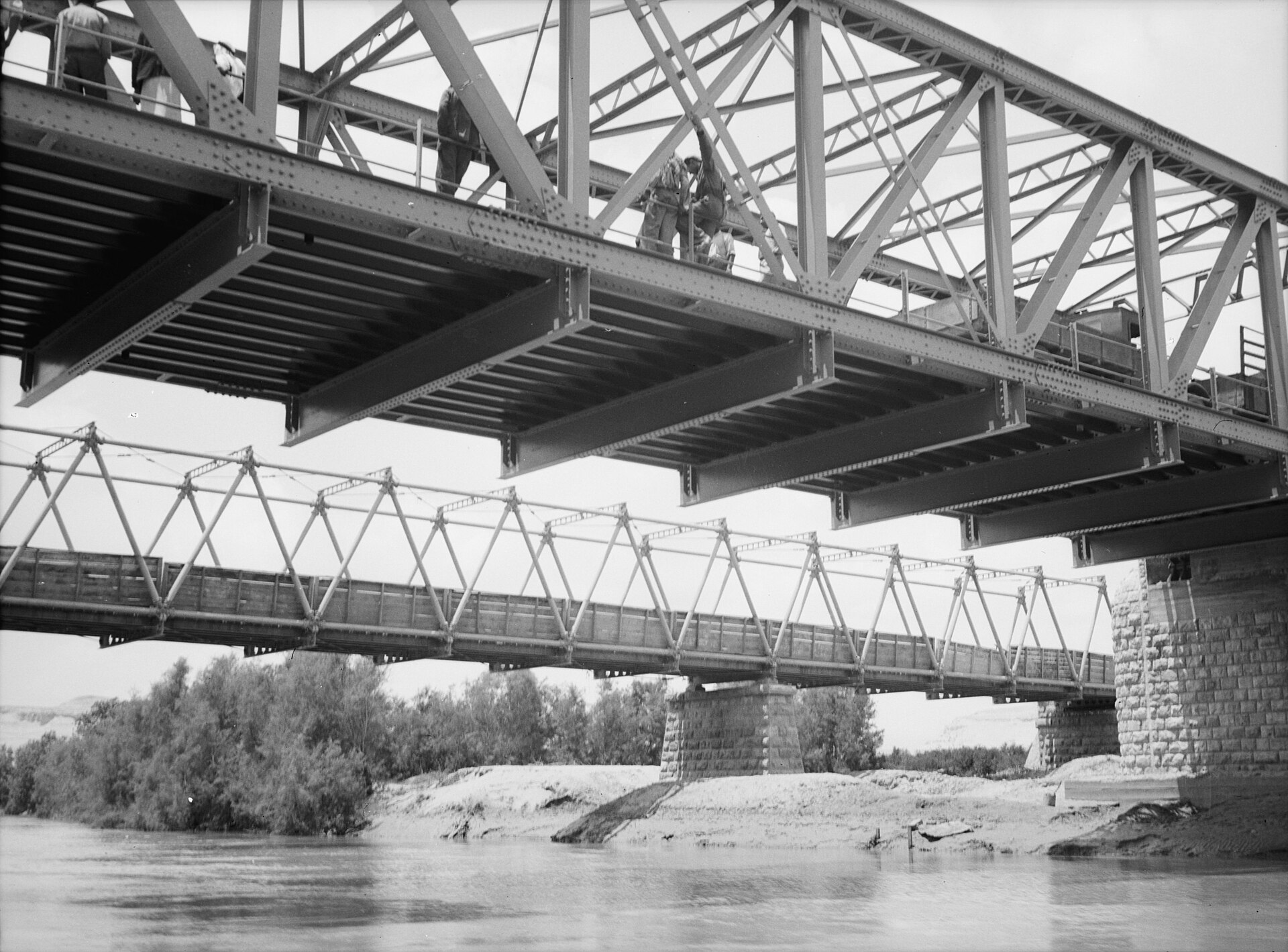 Black and white photograph of a newer steel truss bridge passing over an older bridge across the Jordan River, with trees along the banks