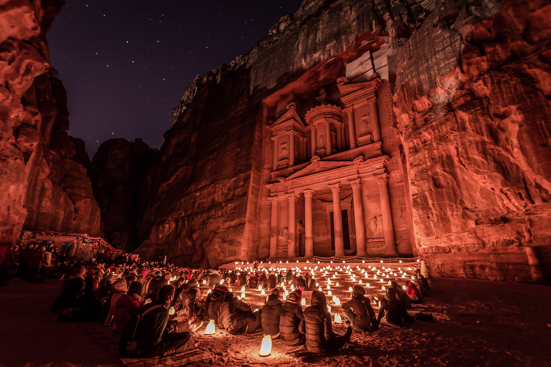 The Treasury facade at Petra lit red at night, with rows of candle lanterns and seated visitors on the ground in front