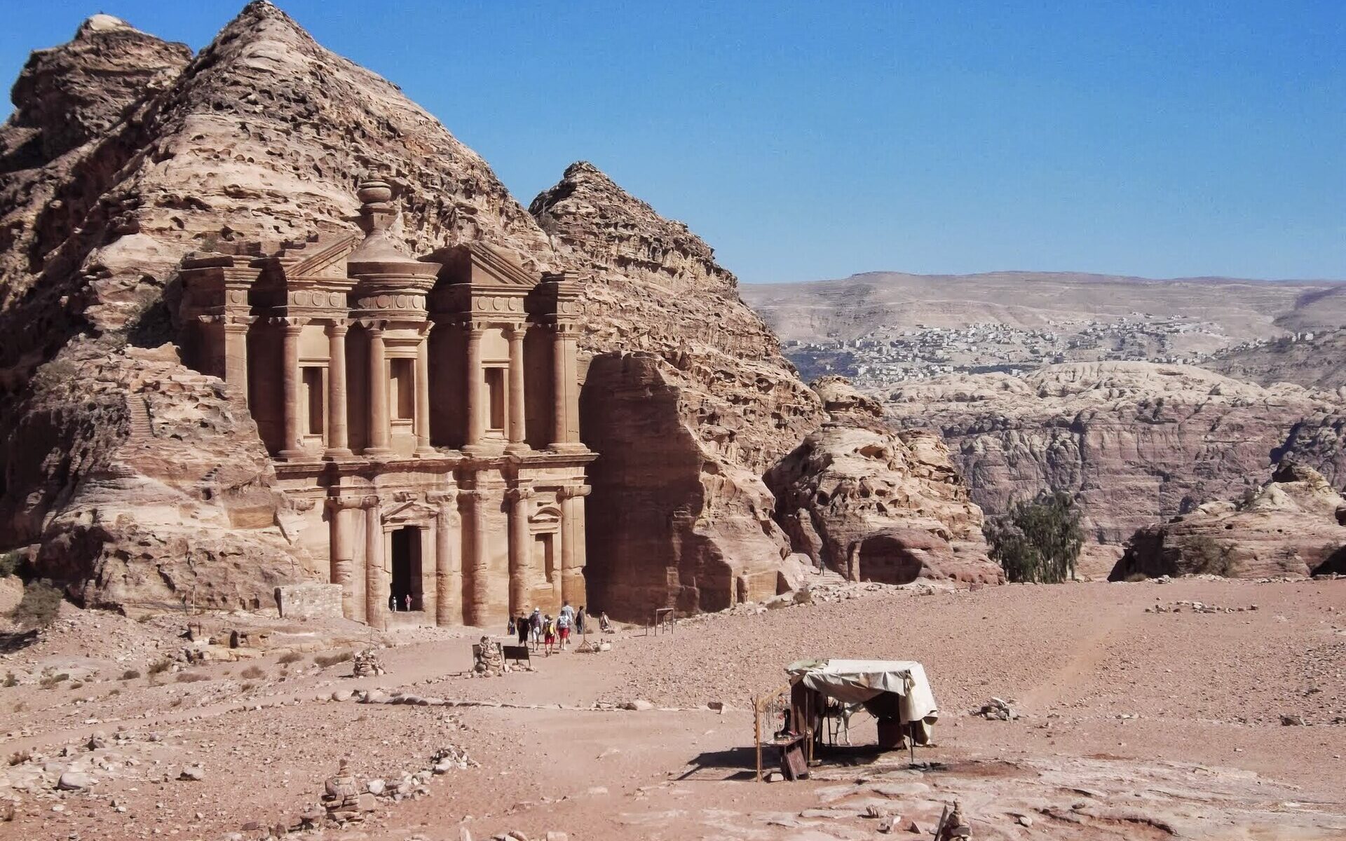 The carved sandstone facade of Ad-Deir, the Monastery, at Petra, set into a rocky hillside under a clear blue sky with a small stall in the foreground
