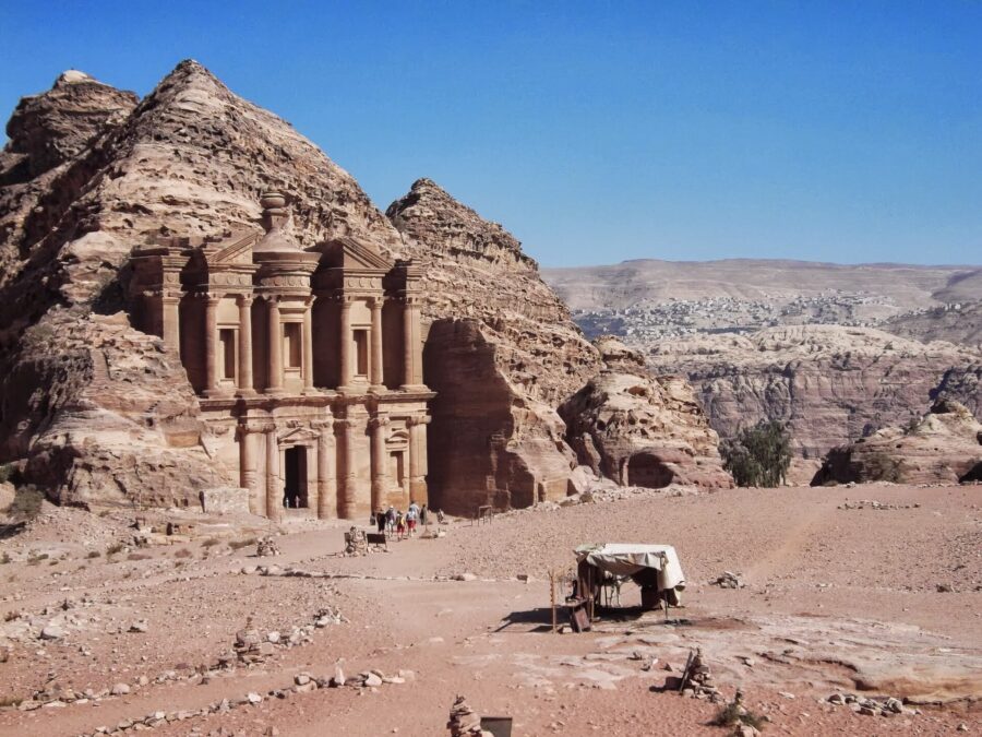 The carved sandstone facade of Ad-Deir, the Monastery, at Petra, set into a rocky hillside under a clear blue sky with a small stall in the foreground