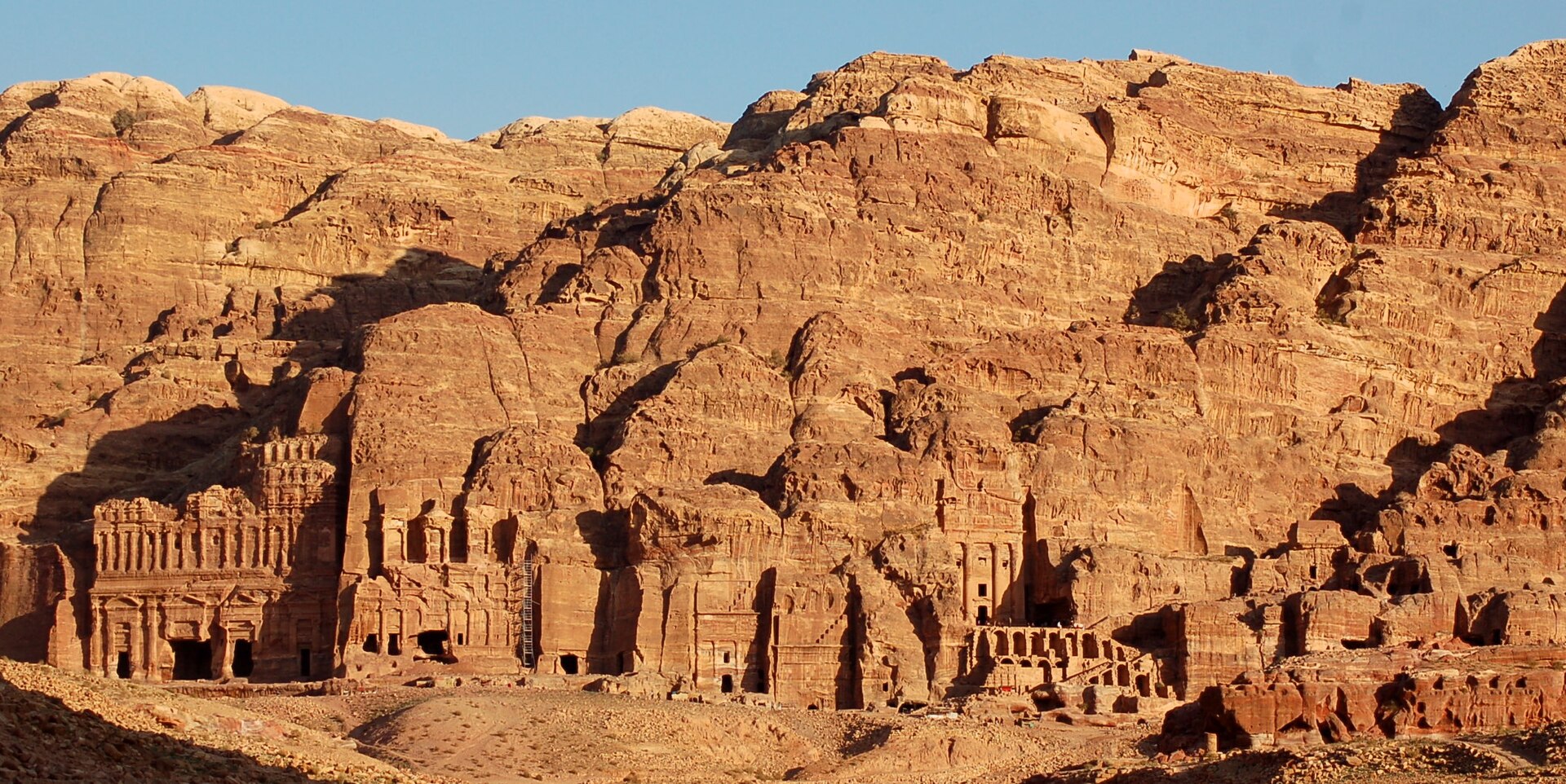 A row of large tomb facades carved into an orange sandstone cliff at Petra, lit by low sunlight