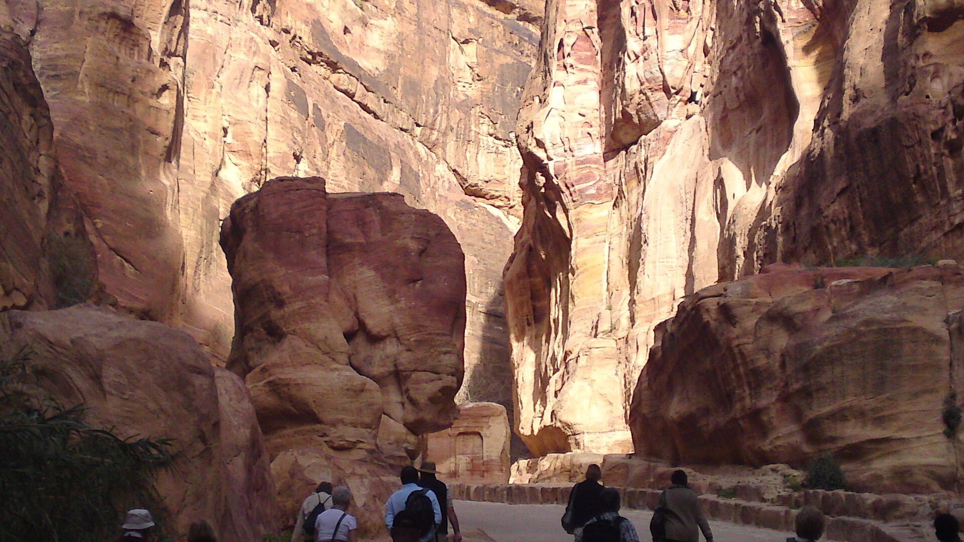 Visitors walking through the narrow Siq gorge at Petra between towering pink and orange sandstone walls, past a large rock formation