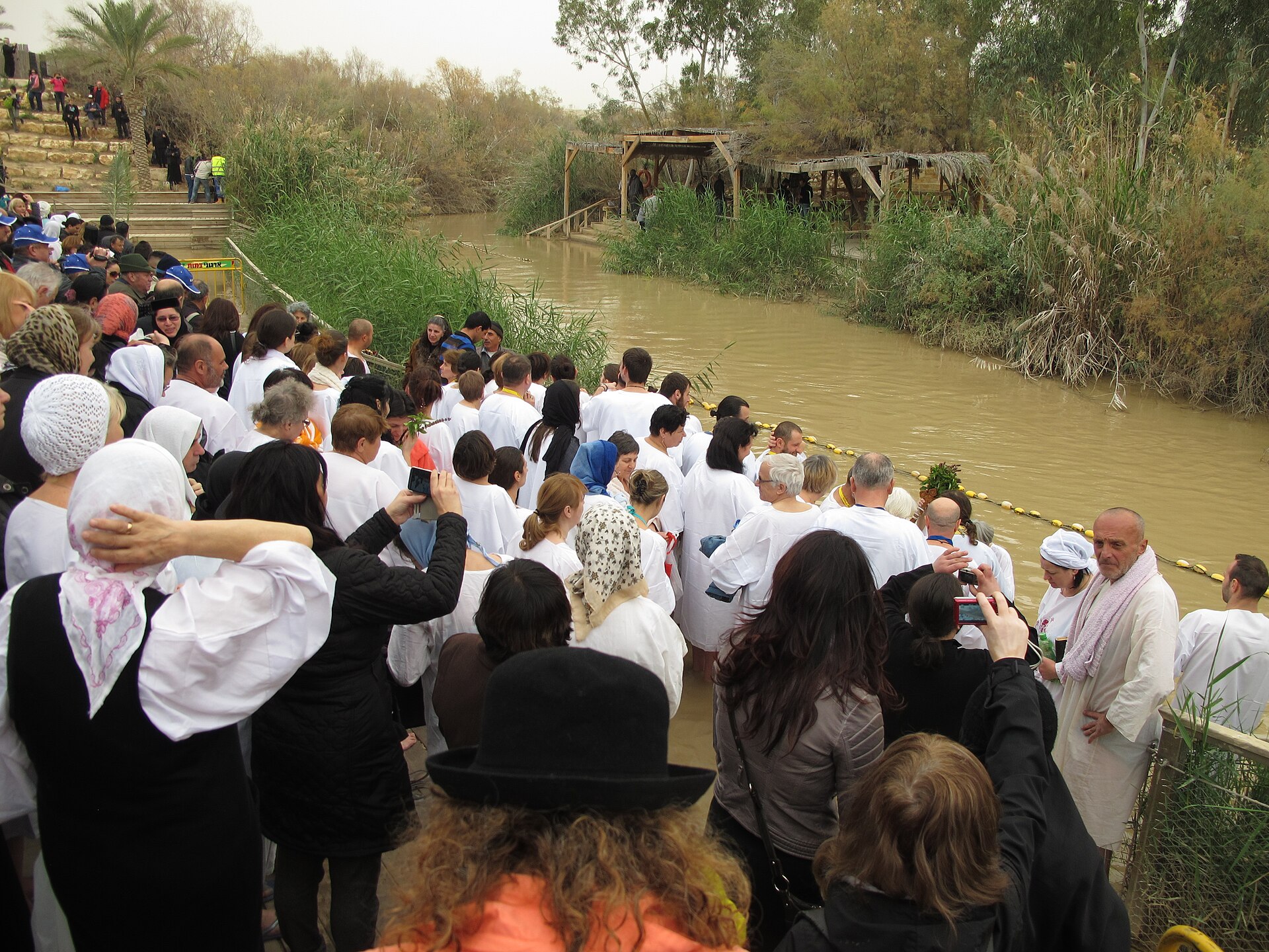 A crowd of pilgrims, many in white robes and head coverings, gathered on the bank of the brown Jordan River among reeds and trees