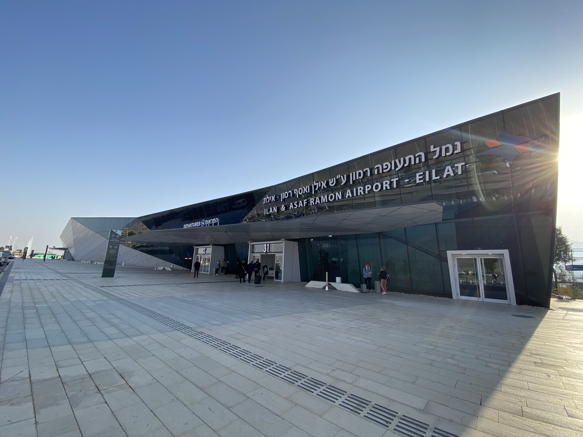 The angular dark glass terminal building of Ramon Airport near Eilat, with its name in Hebrew and English along the roofline and a wide paved forecourt
