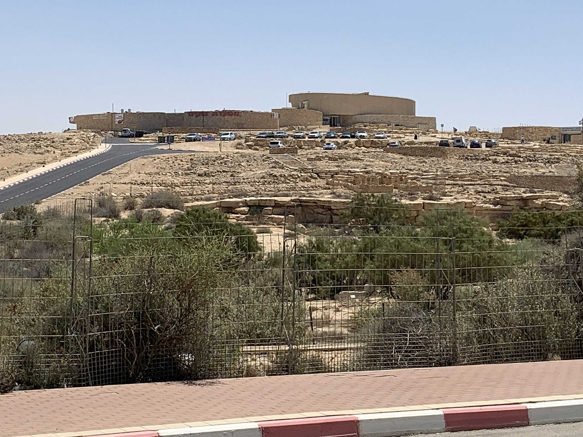 A round sand-coloured building on the edge of a rocky plateau, with a car park in front and desert scrub in the foreground