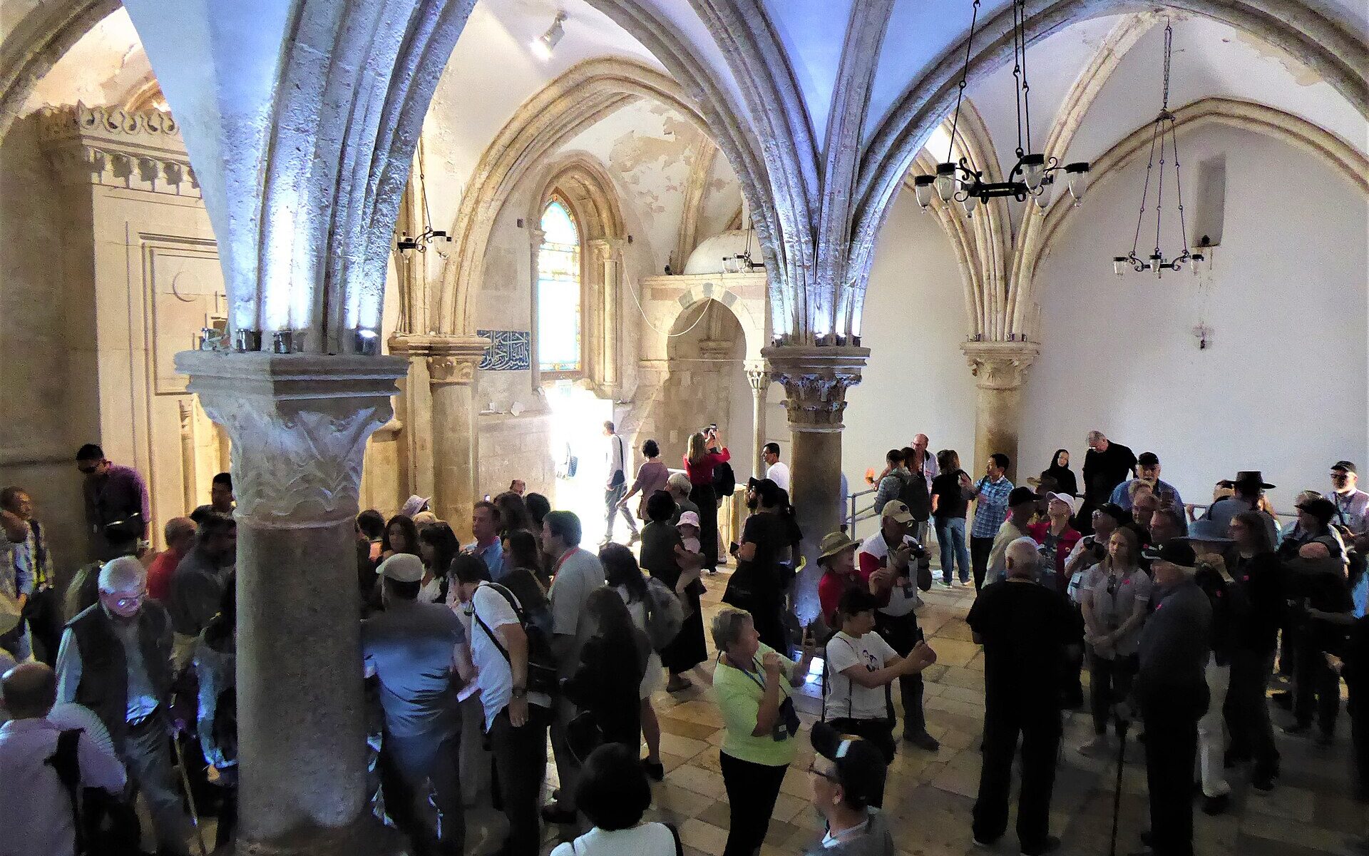 A crowded vaulted Gothic hall with slender columns and ribbed pointed arches, full of visitors standing in groups