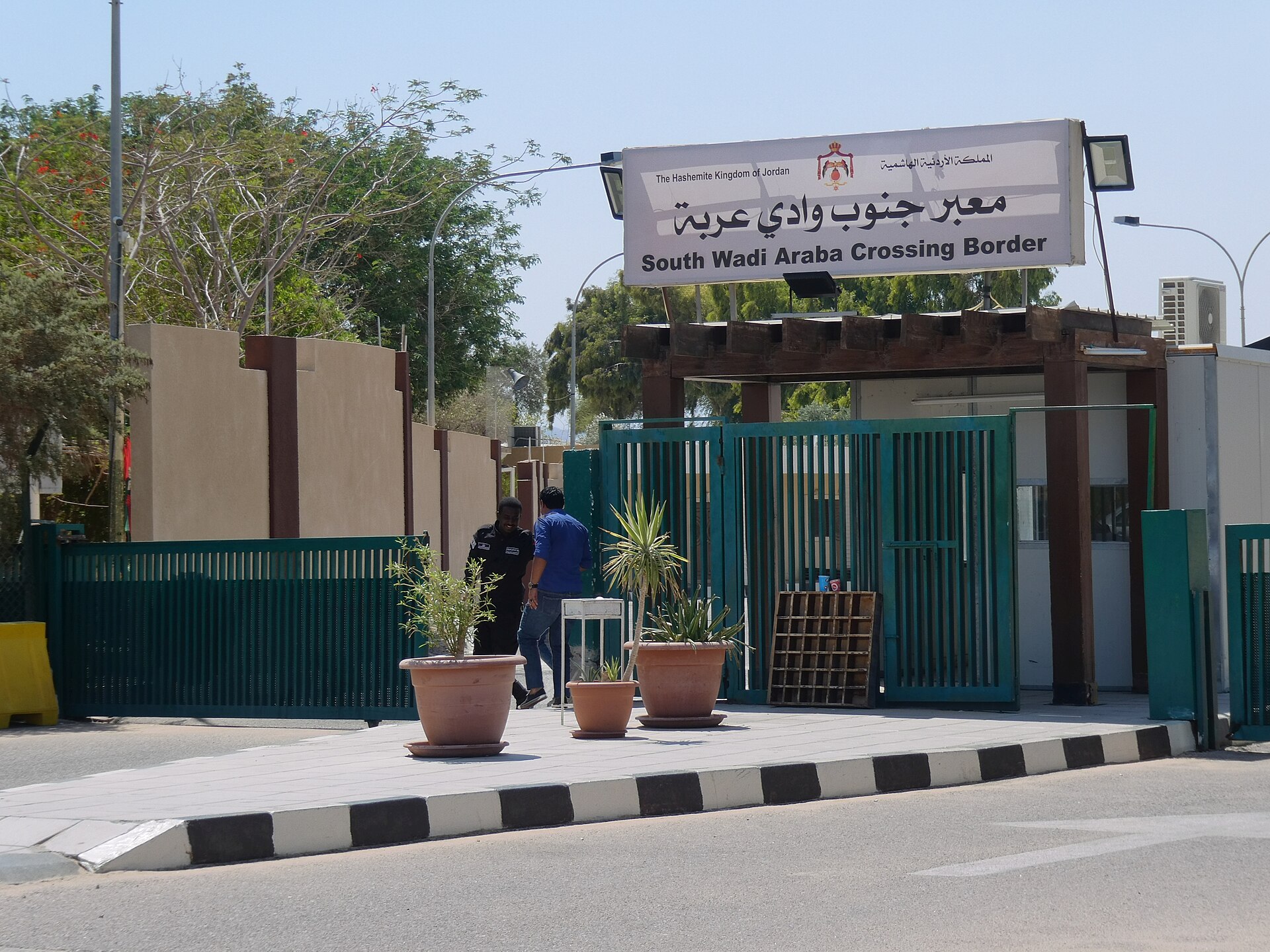The entrance gate of the South Wadi Araba crossing on the Jordanian side, with a bilingual Arabic and English sign above green railings and potted plants