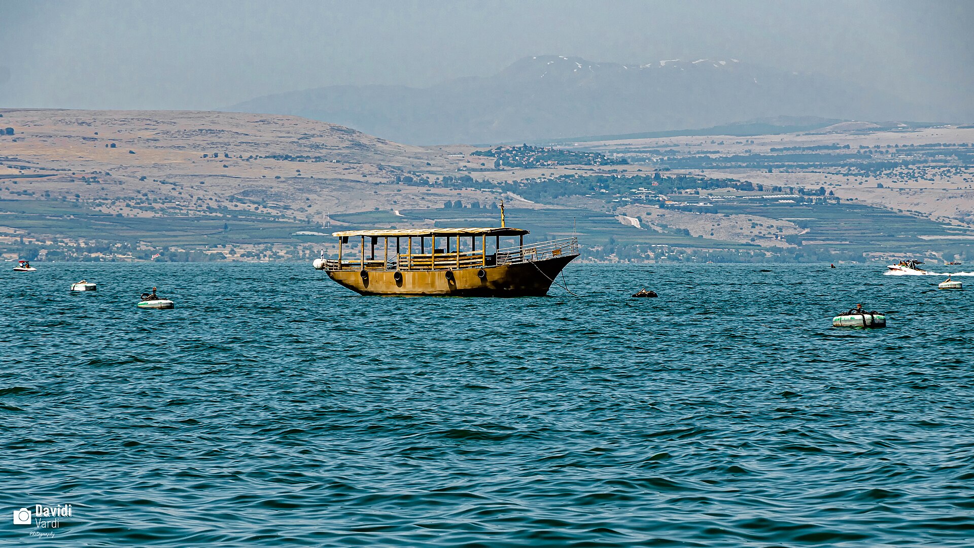 A wooden-style tour boat at anchor on the blue Sea of Galilee with hills on the far shore