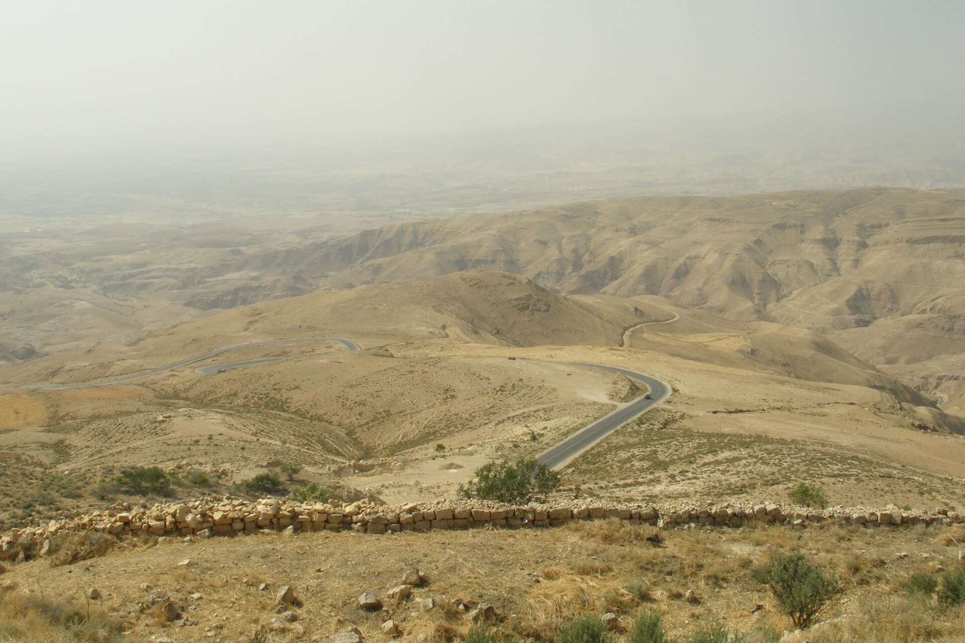 A wide view over dry, folded hills descending towards a hazy valley from a high viewpoint, with a road winding down the slope