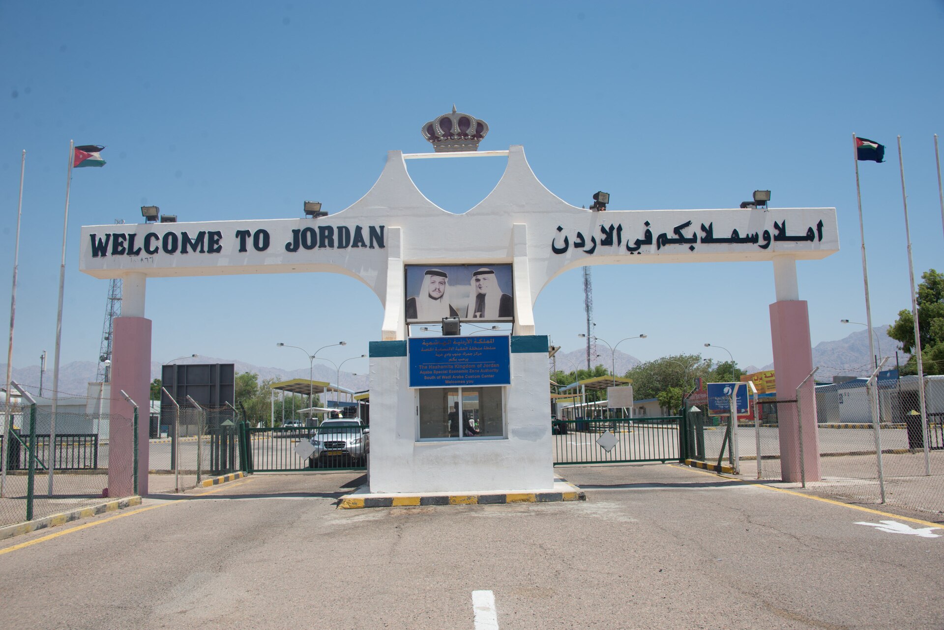 A white arched gateway with a royal crown and portrait, reading Welcome to Jordan in English and Arabic, over the road at a border post with flags on either side